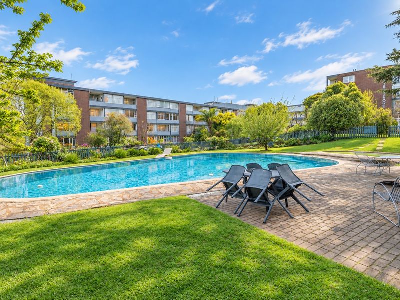 A well-maintained communal swimming pool area featuring stone paving, lush lawn, and established garden landscaping within an apartment complex.