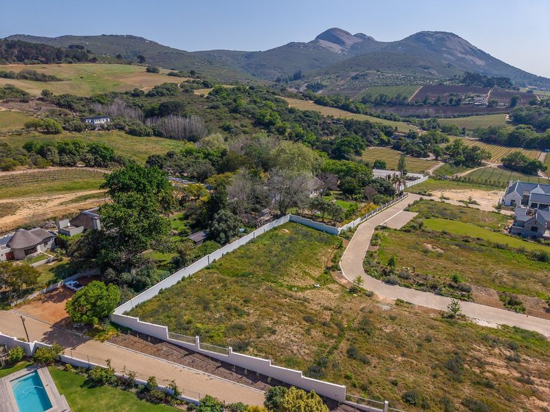 Aerial view of a vacant residential plot featuring perimeter walling and paved road access, set within a scenic landscape with mountain views.