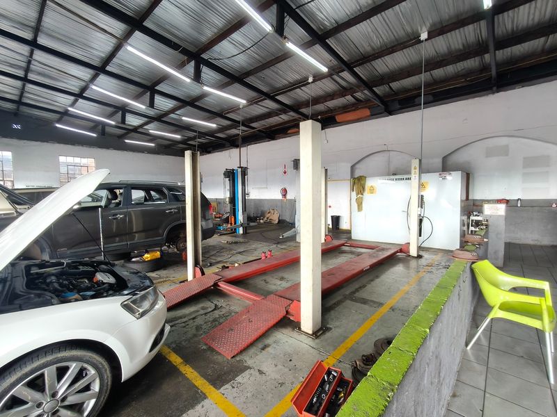 A functional automotive workshop featuring a vehicle hoist, exposed industrial ceiling, and a dedicated spray booth area with concrete flooring.