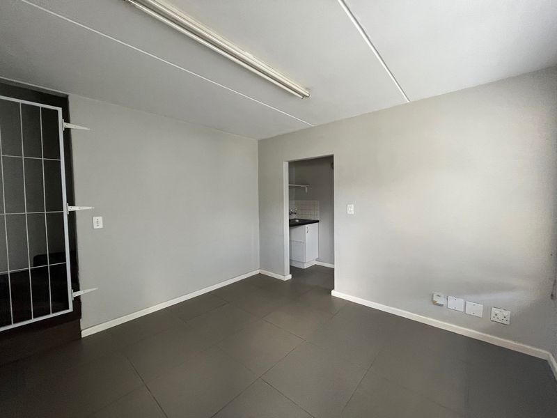 An open-plan industrial space featuring tiled flooring, a fluorescent light fixture, and a small adjoining kitchenette area with a security gate at the entrance.