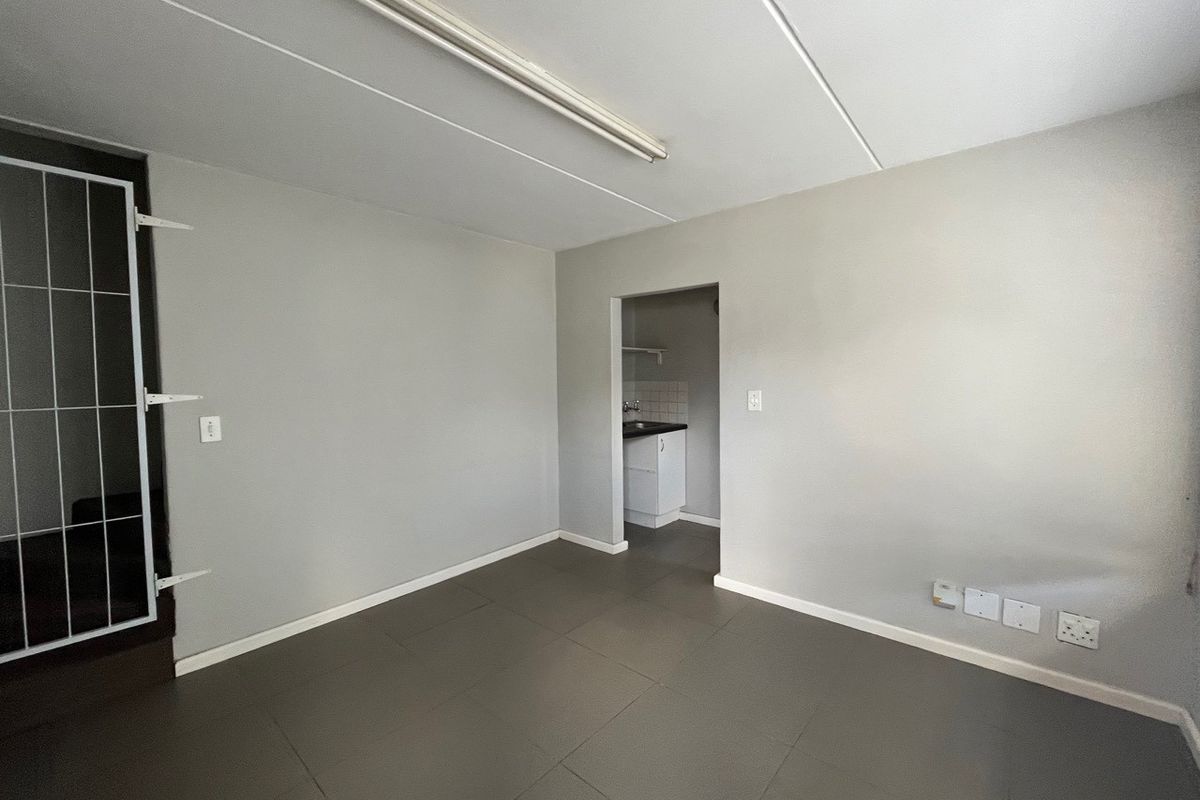An open-plan industrial space featuring tiled flooring, a fluorescent light fixture, and a small adjoining kitchenette area with a security gate at the entrance.