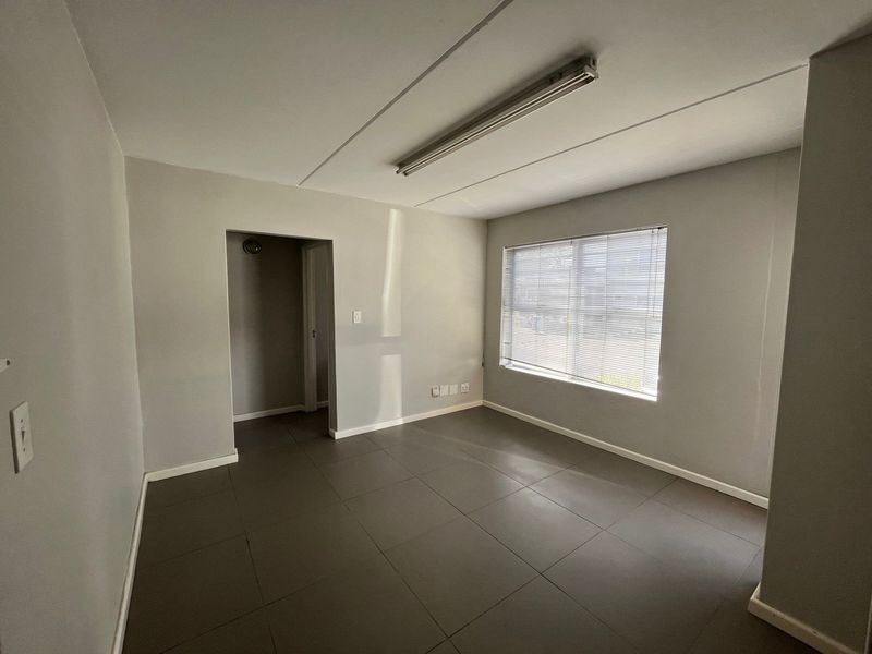 A clean, empty office space featuring neutral grey walls, tiled flooring, and a large window with fitted venetian blinds.