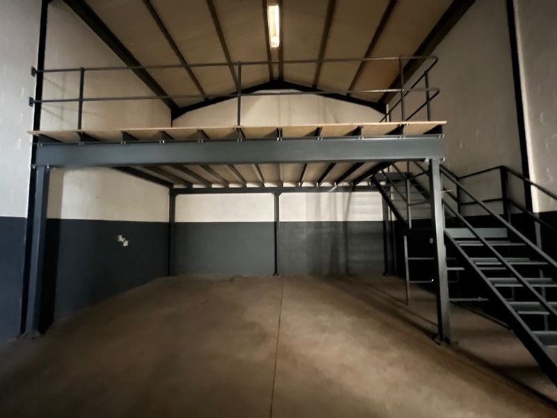 Spacious industrial utility area featuring a mezzanine level with steel staircase, high ceilings, and durable concrete flooring, suitable for storage or workshop use.