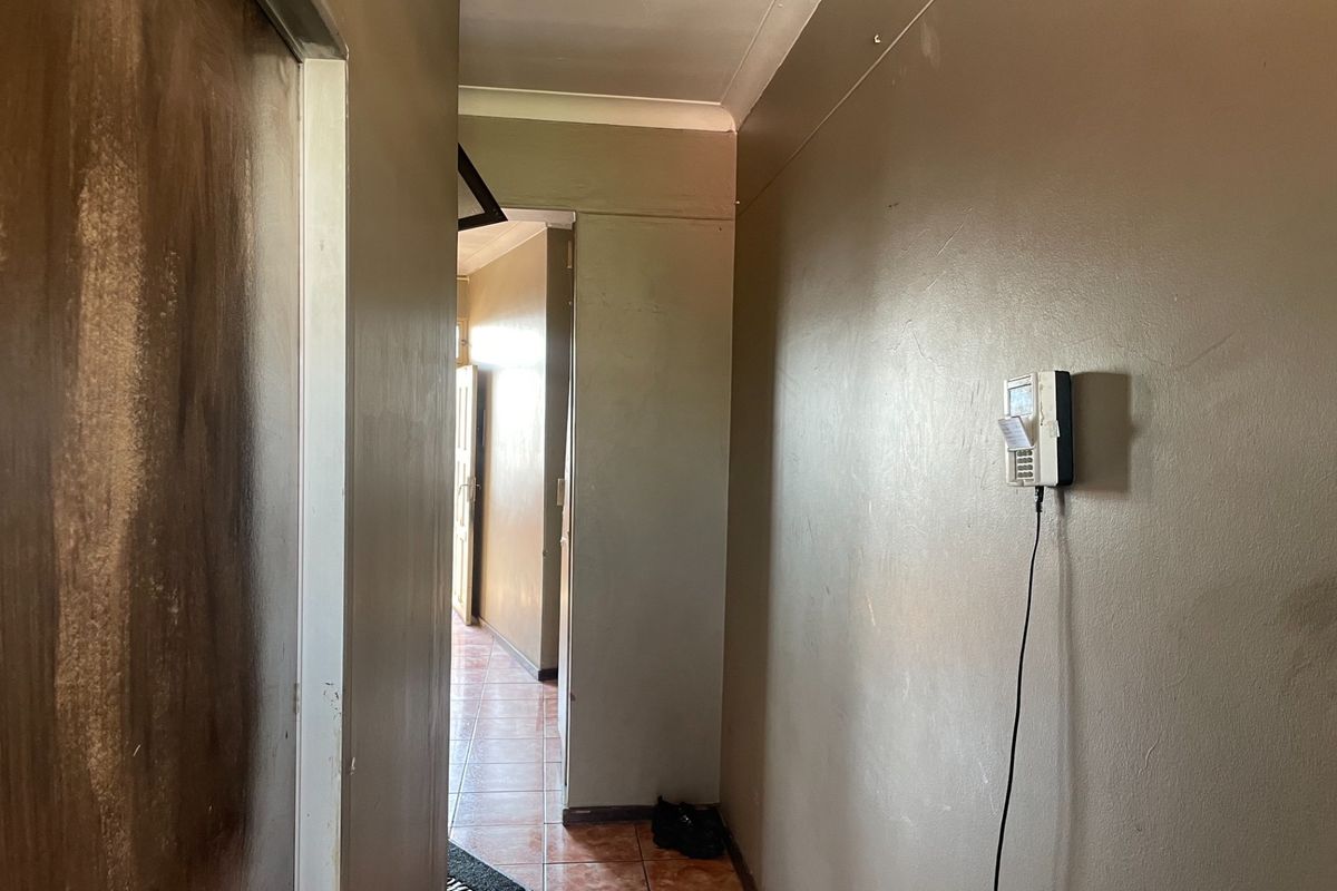 A narrow hallway featuring tiled flooring, coved ceilings, and a wall-mounted intercom system, showing signs of wear on the walls and door frames.