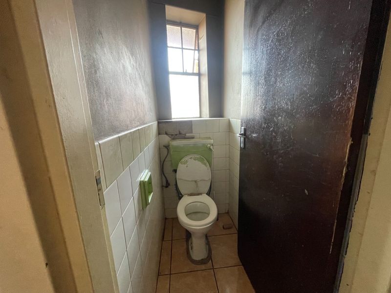 A small, dated bathroom featuring a toilet, ceramic tiling, and a window. The space requires renovation and cosmetic updates to improve its condition.