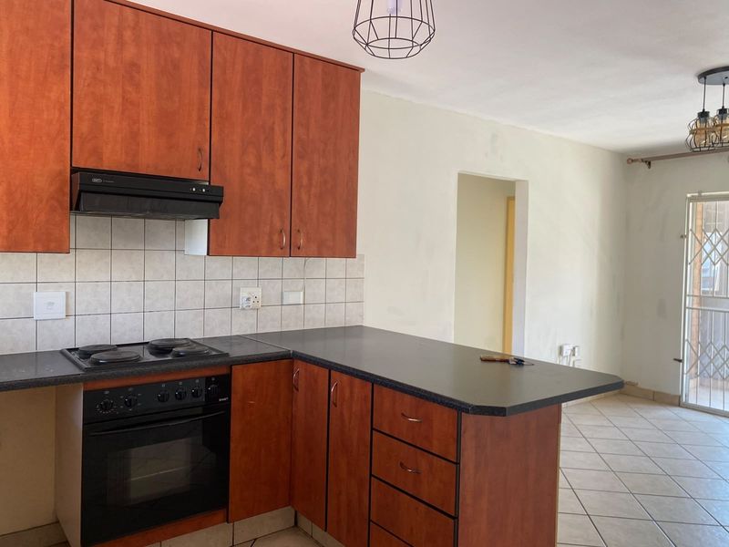 A functional kitchen featuring wood-finish cabinetry, dark laminate countertops, and built-in appliances, opening into an adjacent living area with tiled flooring.