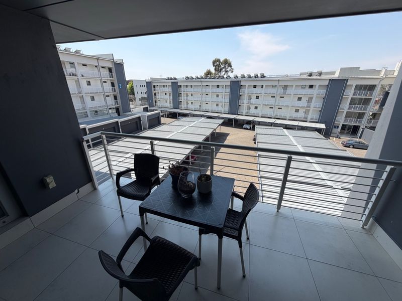 A modern apartment balcony featuring tiled flooring and a metal balustrade, overlooking the complex parking area.