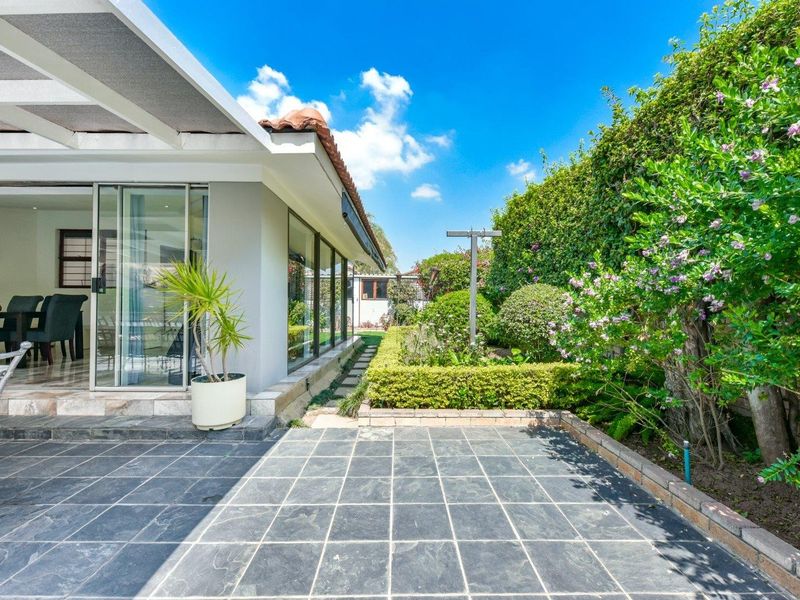 A well-maintained tiled patio area featuring sliding glass doors leading to the interior, complemented by a lush, landscaped garden with mature hedges.
