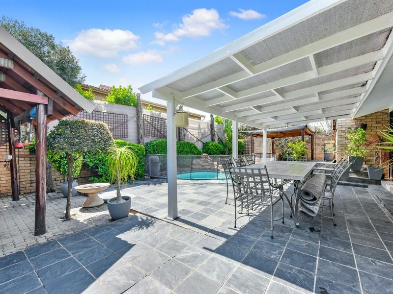 A spacious covered patio area featuring stone tile flooring, overlooking a private swimming pool and landscaped garden with brick boundary walls.