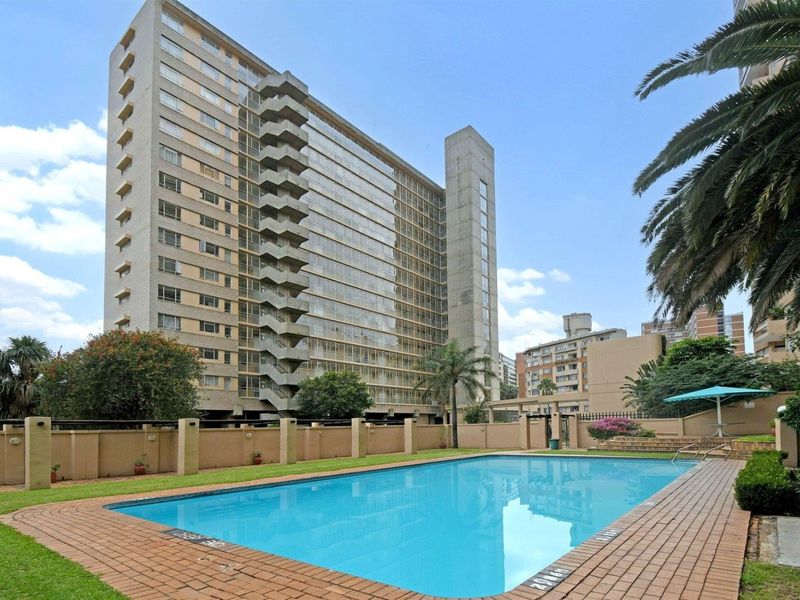 A well-maintained communal swimming pool area featuring brick paving, landscaped garden surrounds, and a perimeter wall, set against a high-rise apartment building.