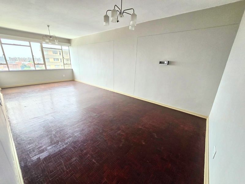 A spacious, empty living area featuring original parquet flooring, large windows providing natural light, and neutral wall finishes.