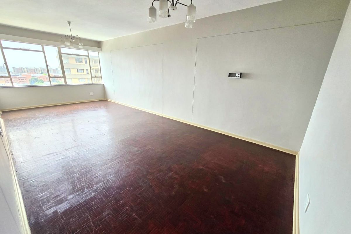 A spacious, empty living area featuring original parquet flooring, large windows providing natural light, and neutral wall finishes.