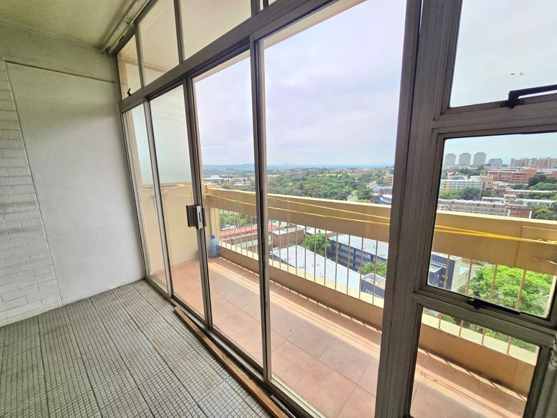 An enclosed balcony featuring sliding glass doors, tiled flooring, and a metal balustrade, offering an elevated urban view.