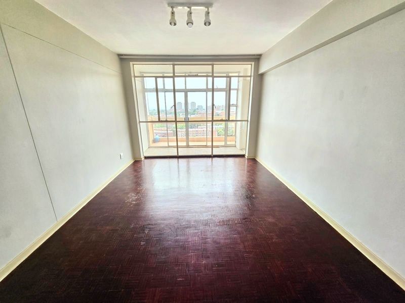 An empty living room featuring original parquet flooring, large glass sliding doors leading to a balcony, and neutral wall finishes.