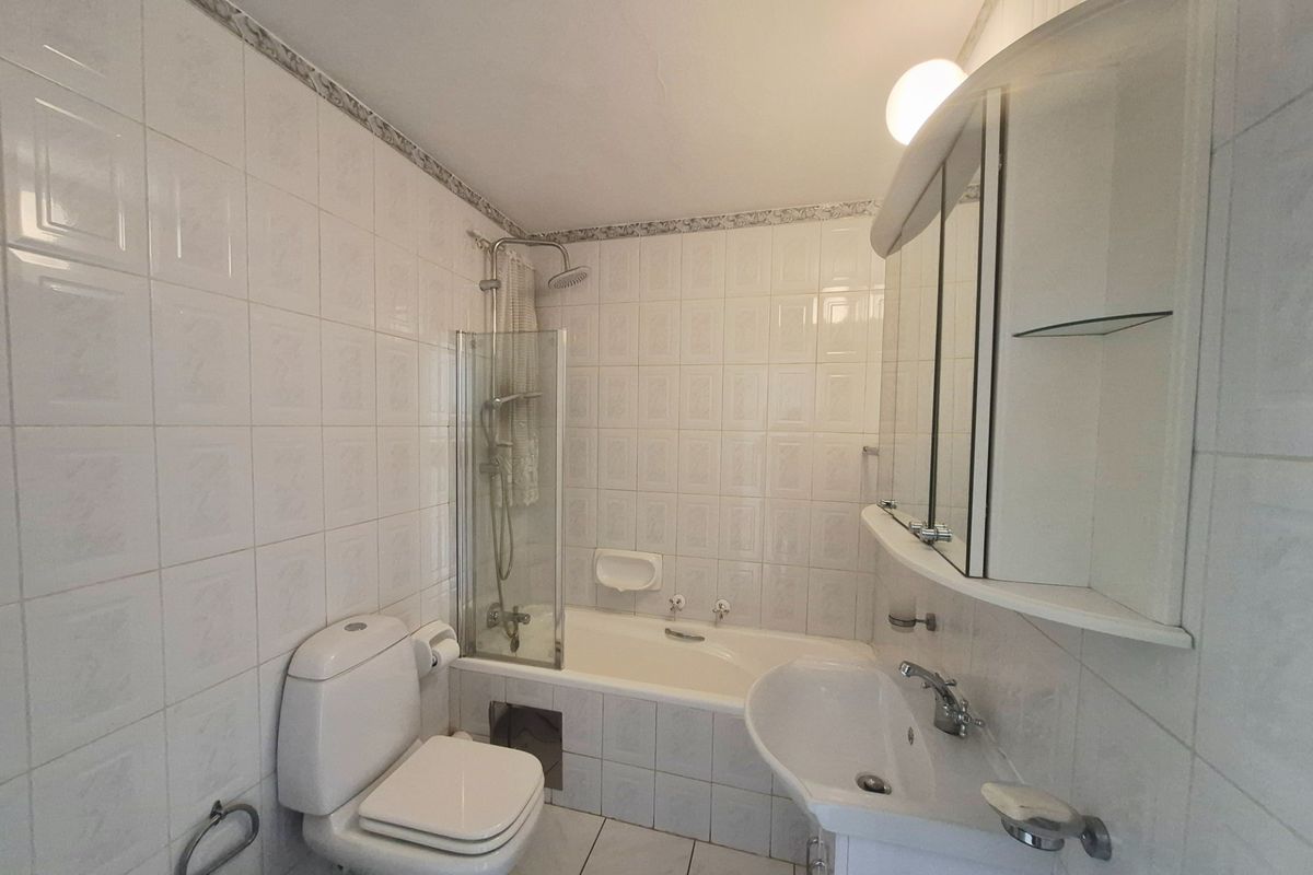 A functional bathroom featuring a bathtub with shower attachment, pedestal sink, and mirrored cabinetry, finished with classic white ceramic wall and floor tiles.