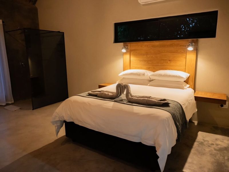 A modern bedroom featuring polished concrete floors, a custom wooden headboard with integrated lighting, floating bedside shelves, and an en-suite glass shower enclosure.