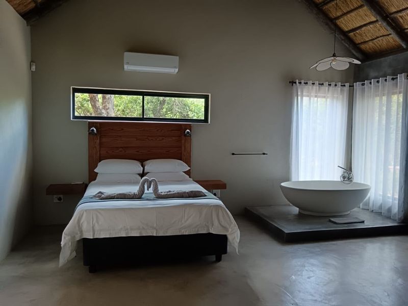 Spacious bedroom featuring a thatched roof with exposed rafters, polished concrete floors, a built-in timber headboard, and a luxurious freestanding bathtub.