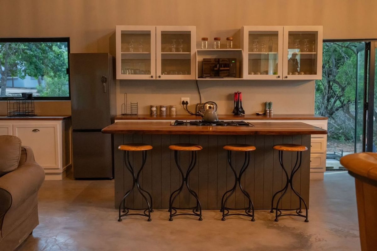 A modern kitchen featuring a central island with a timber countertop, gas hob, ample cabinetry, and polished concrete floors, opening to an outdoor area.