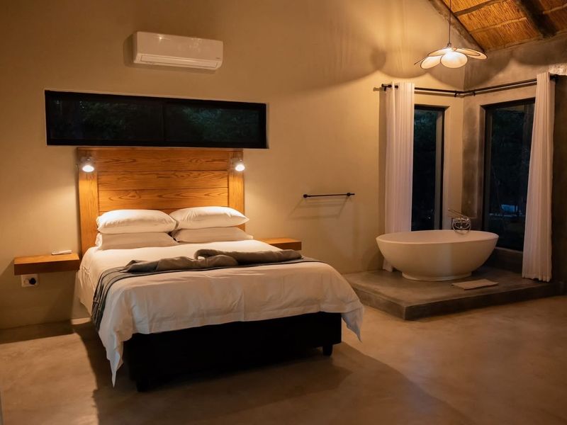 Spacious bedroom featuring a thatched ceiling, polished concrete flooring, a built-in wooden headboard, and a luxurious freestanding bathtub on a raised platform.