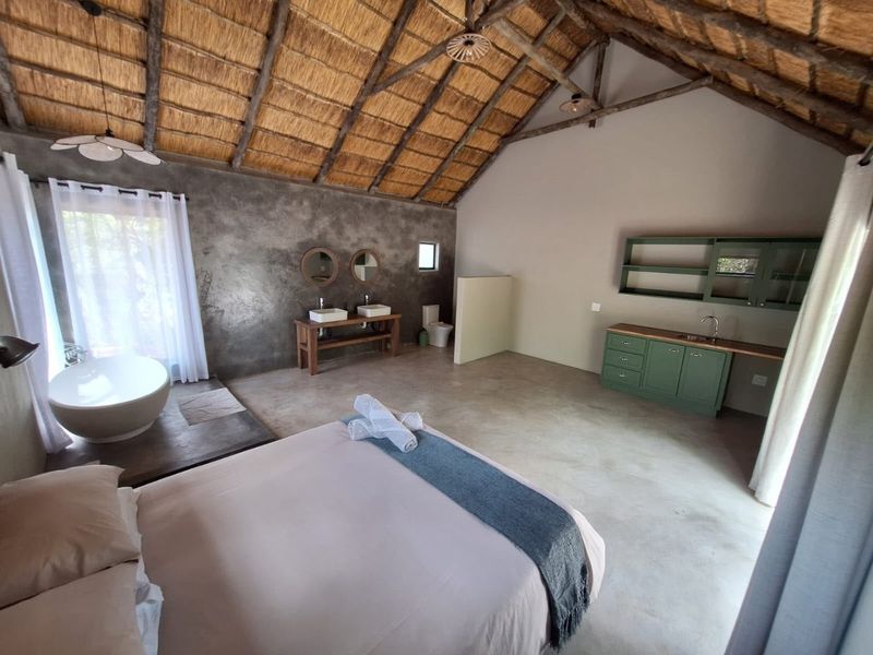 Spacious bedroom featuring a high thatched roof with exposed trusses, concrete flooring, a freestanding bathtub, double vanity, and integrated cabinetry.