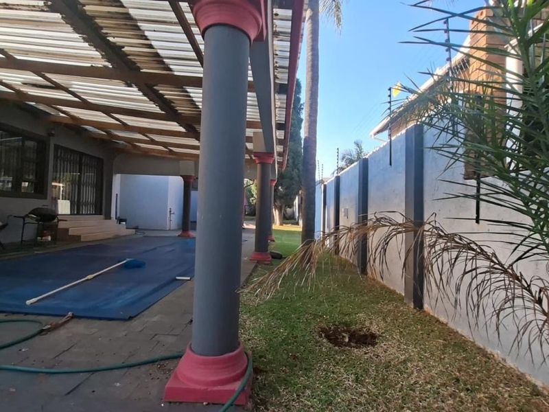 A covered outdoor patio area featuring concrete columns, paved flooring, and a perimeter wall with electric fencing, overlooking a small garden space.