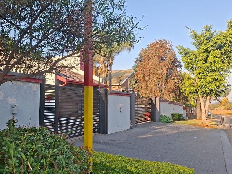 Street view of the property featuring a paved driveway, boundary wall with metal gates, and established trees along the roadside.