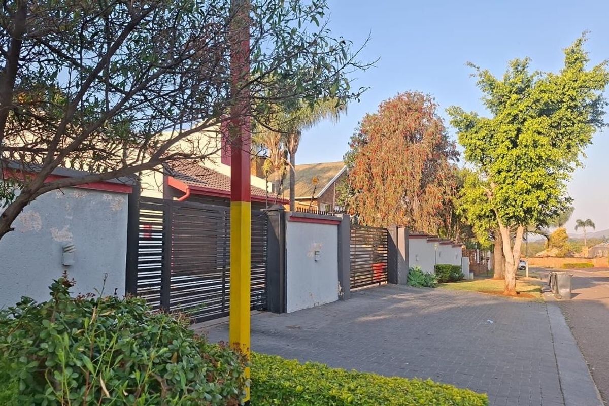 Street view of the property featuring a paved driveway, boundary wall with metal gates, and established trees along the roadside.