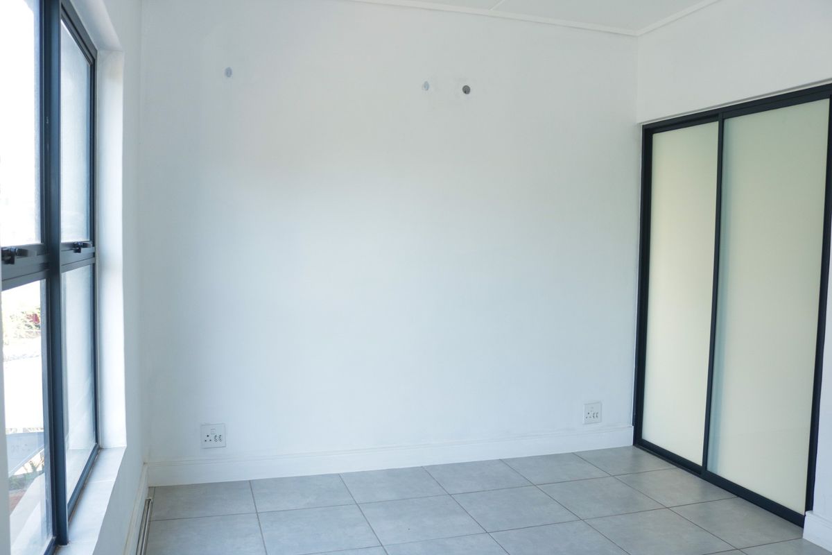 A bright, empty bedroom featuring neutral tiled flooring, a modern built-in sliding door wardrobe, and large windows providing natural light.
