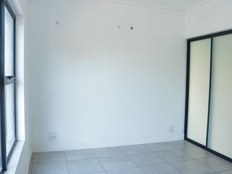A bright, empty bedroom featuring neutral tiled flooring, a modern built-in sliding door wardrobe, and large windows providing natural light.