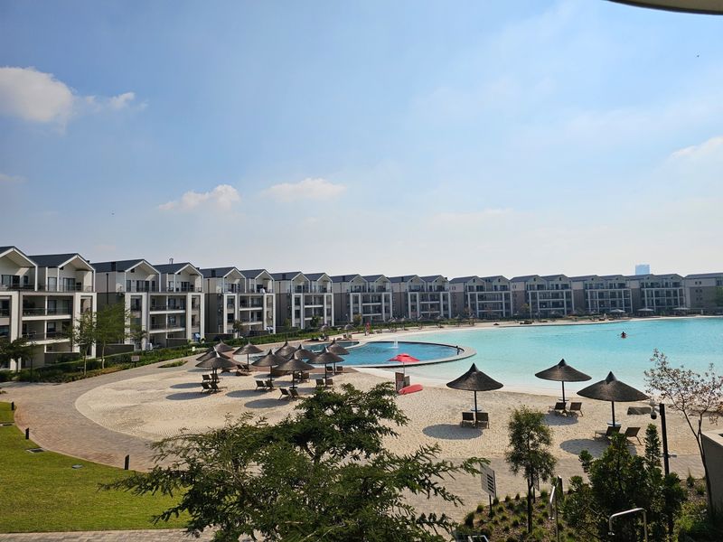 A scenic view of a large communal swimming pool with a sandy beach area, surrounded by modern apartment buildings and landscaped grounds.