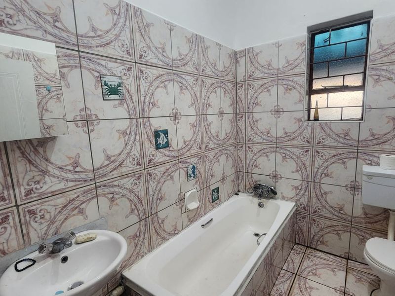 A dated bathroom featuring full-height patterned ceramic wall tiles, a standard bathtub, pedestal sink, and toilet, with a window providing natural light.