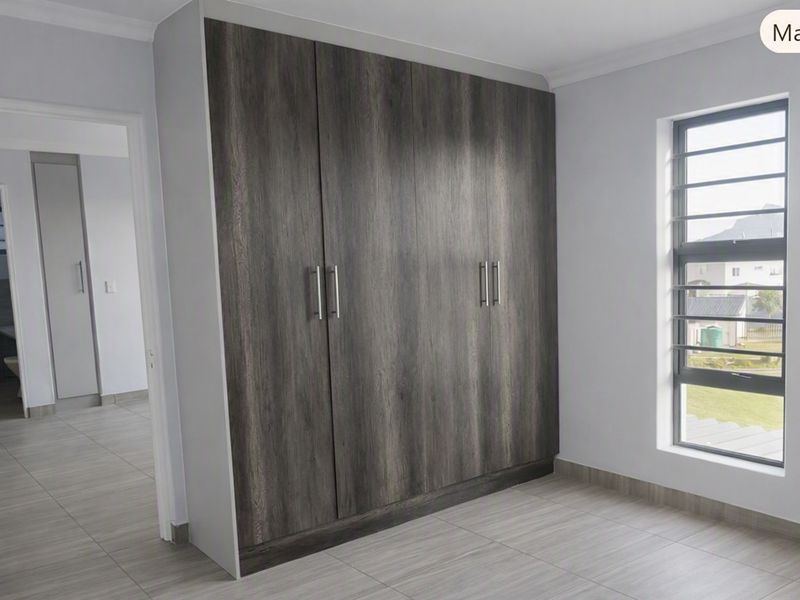 A bright, modern bedroom featuring neutral tiled flooring, built-in dark wood-grain cupboards, and a louvered window providing natural light.