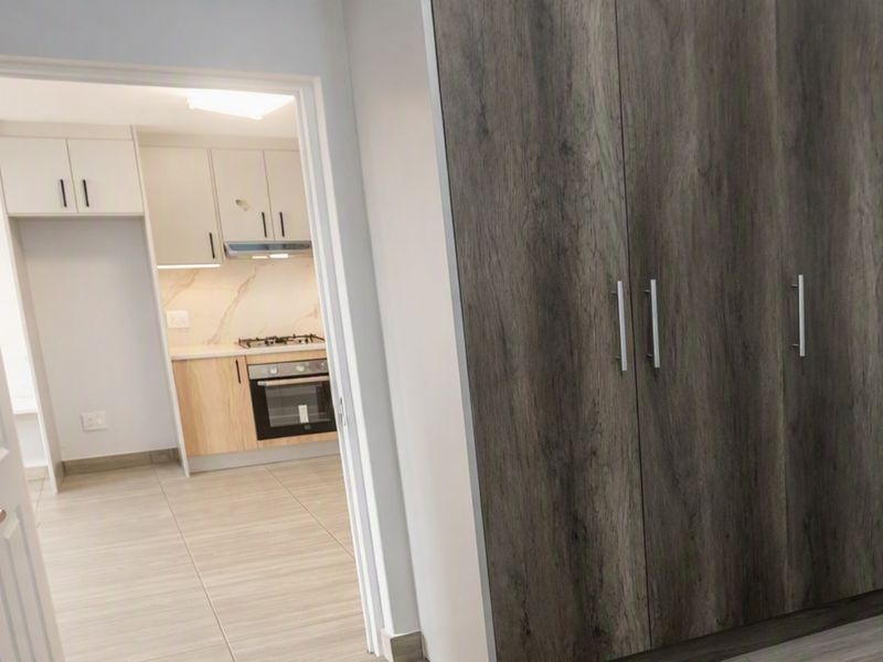 A clean bedroom featuring modern wood-look built-in cupboards and tiled flooring, with a view through the doorway into the adjacent kitchen area.