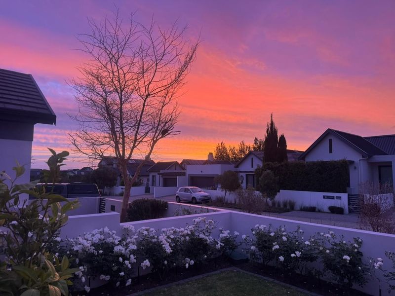 A picturesque front exterior view of a modern residential property featuring a well-maintained garden and boundary wall under a vibrant sunset sky.