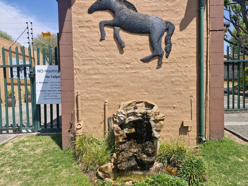 Entrance area of the apartment complex featuring a stone water feature, decorative metal horse sculpture, and secure gated access with electric fencing.