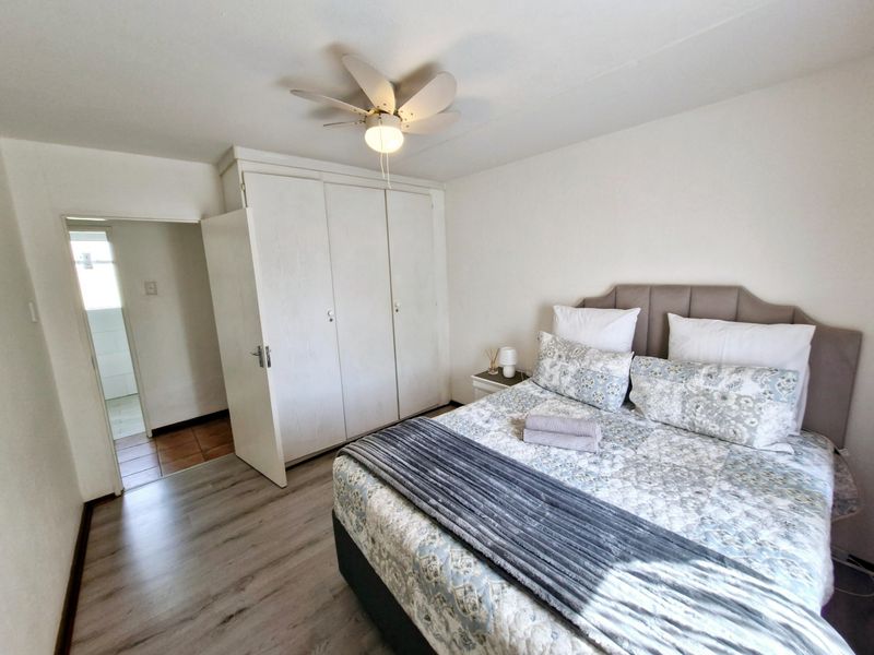 A well-lit bedroom featuring built-in cupboards, a ceiling fan, and light-toned laminate flooring, with direct access to an adjoining bathroom.