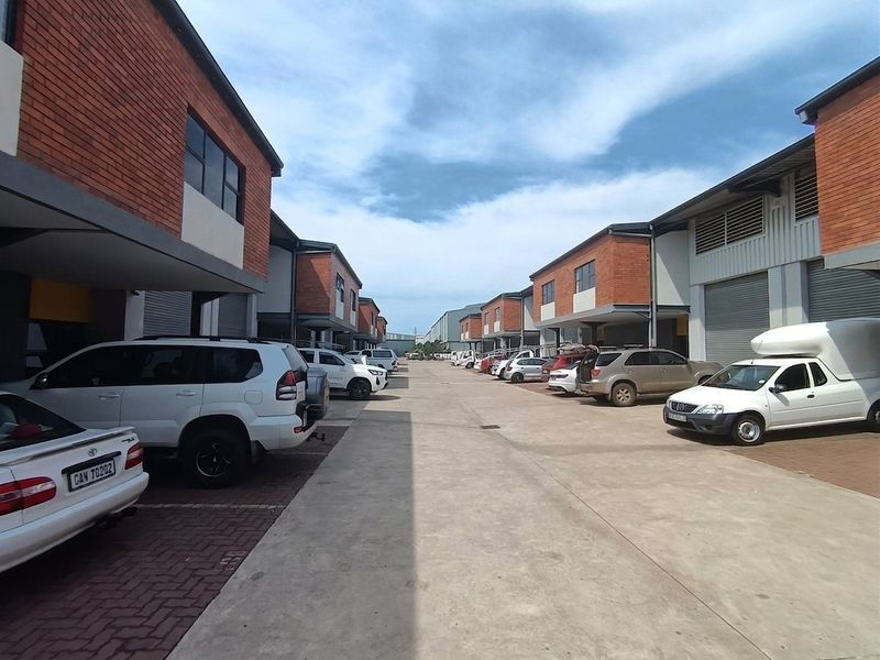 An industrial complex exterior featuring a central concrete driveway, paved parking bays, and multiple units with roller shutter doors and brick facades.