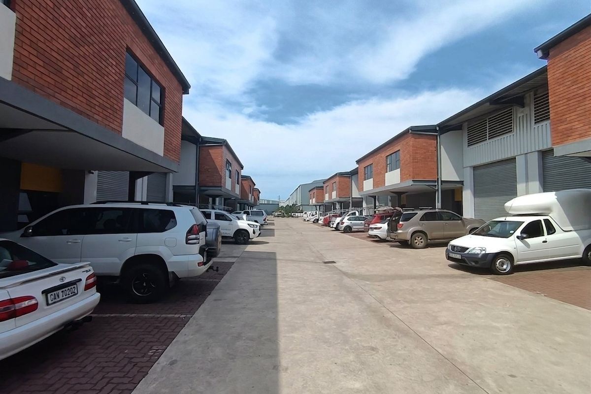 An industrial complex exterior featuring a central concrete driveway, paved parking bays, and multiple units with roller shutter doors and brick facades.