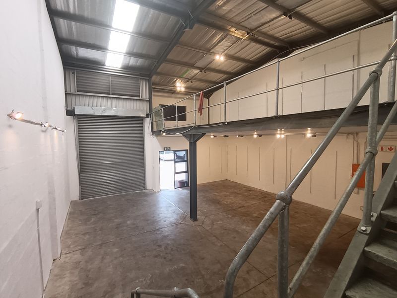 Spacious industrial warehouse unit featuring a high-clearance ceiling, roller shutter door access, and a functional mezzanine level with steel staircase and railings.