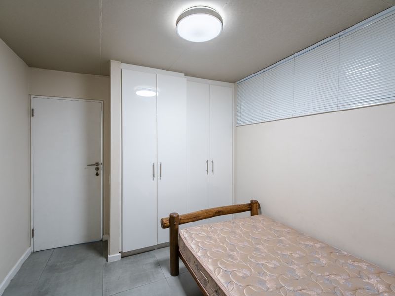 A functional bedroom featuring built-in white cupboards, tiled flooring, and venetian blinds, providing a clean and neutral space.
