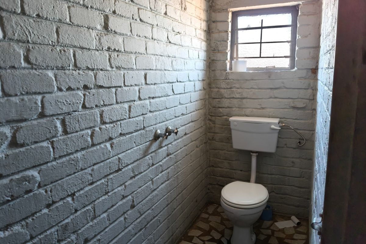 A small, utilitarian bathroom featuring exposed painted brick walls, a standard toilet, and a small window with security bars.