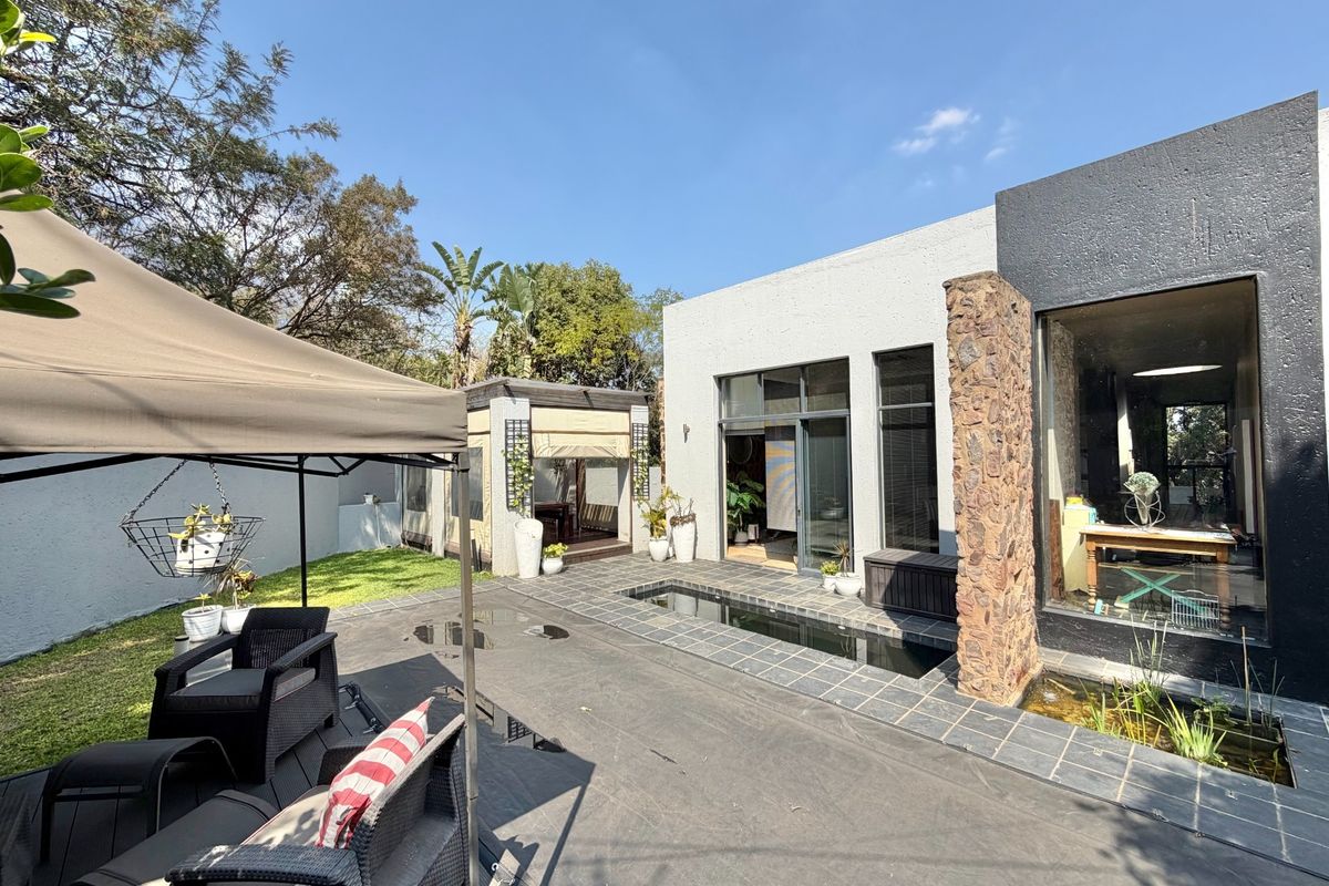 A modern rear garden area featuring a tiled patio, a stone-clad pillar, a built-in water feature, and a covered gazebo structure.