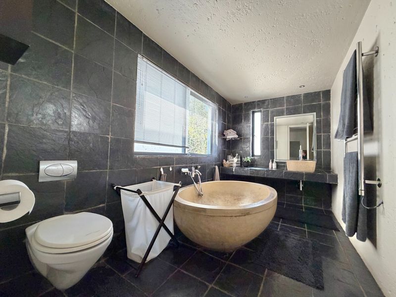 Modern bathroom featuring dark slate tiling, a unique stone freestanding bathtub, wall-mounted toilet, and a stone vanity countertop with a heated towel rail.