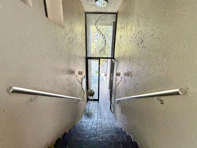 A narrow staircase hallway featuring textured walls, stainless steel handrails, and dark tiled steps leading to a glass door with outdoor access.