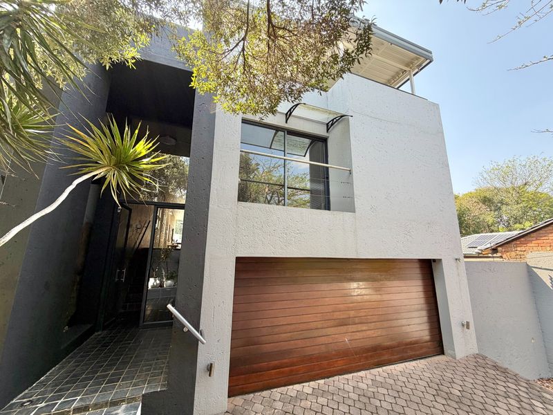 Modern multi-level residential facade featuring a wooden automated garage door, paved driveway, and an upper-level balcony with a glass balustrade.