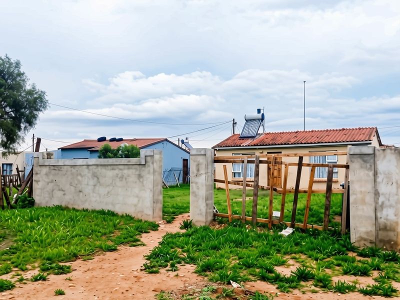 Front view of a residential property featuring a tiled roof, solar water heater, and a partially enclosed yard with a damaged wooden gate.
