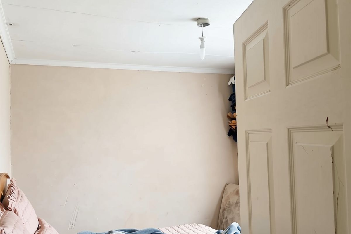 A bedroom featuring a simple ceiling light fixture and a panelled door, showing signs of wear on the walls and ceiling.