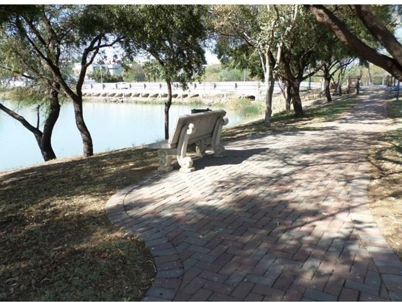 A scenic garden pathway featuring brick paving and a stone bench overlooking a tranquil lake and bridge.