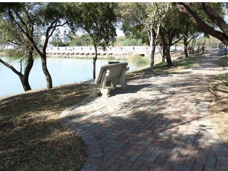 A scenic garden pathway featuring brick paving, mature trees, and a tranquil view overlooking a nearby water feature.