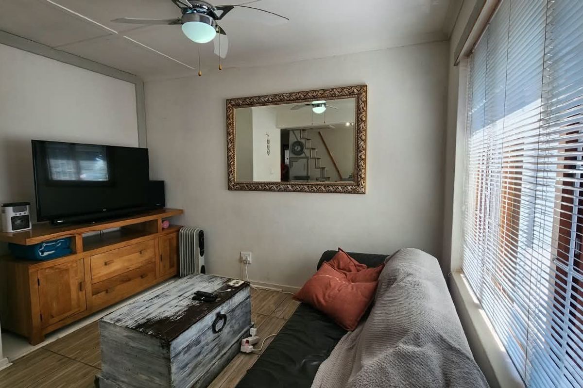 A comfortable living room featuring tiled flooring, a ceiling fan, and large windows fitted with venetian blinds.